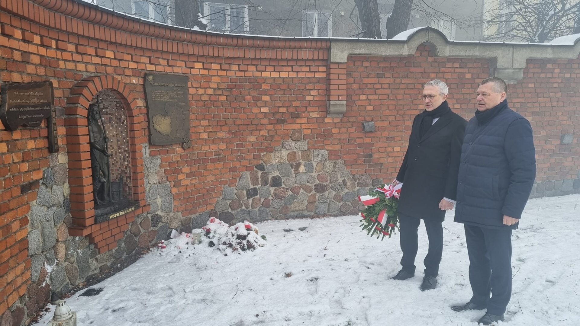 Burmistrz Arkadiusz Ptak i zastępca Andrzej Jędruszek stoją z wiżanką wiatów przed Murem Pamięci, na którymzawieszona jest tablica "Solidarności"