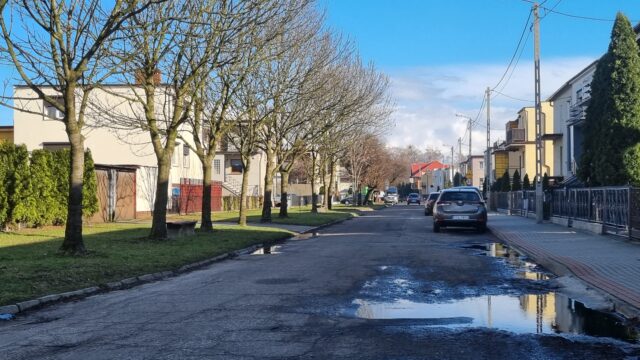 zdjęcie przedstawia ulicę, po prawej stronie stoja budynki i stoi samochód, po lewej rosną drzewa, 