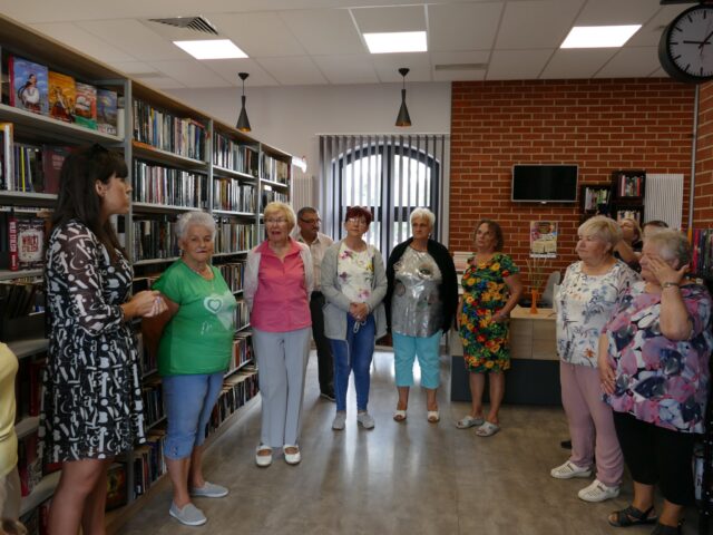 Seniorzy podczas wizyty w Bibliotece Publicznej w ramach półkolonii senioralnych wraz z dyrektor biblioteki