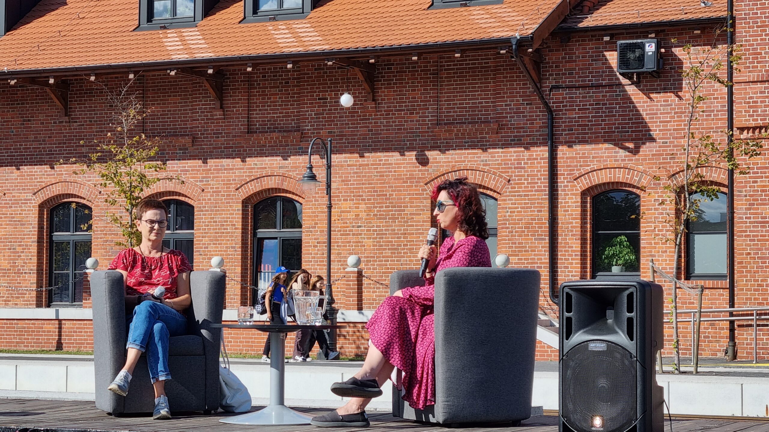 Dwie kobiety siedzące w fotelach i prwadzace rozmowę; w tle Biblioteka Publiczna w Pleszewie