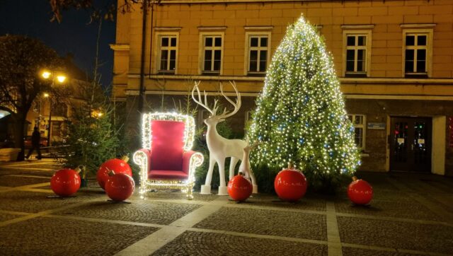 zdjęcie przedstawia świąteczny fotel, duża oświetloną choinkę oraz bombki i renifery wydrukowane na drukarce 3D na pleszewskim Rynku.