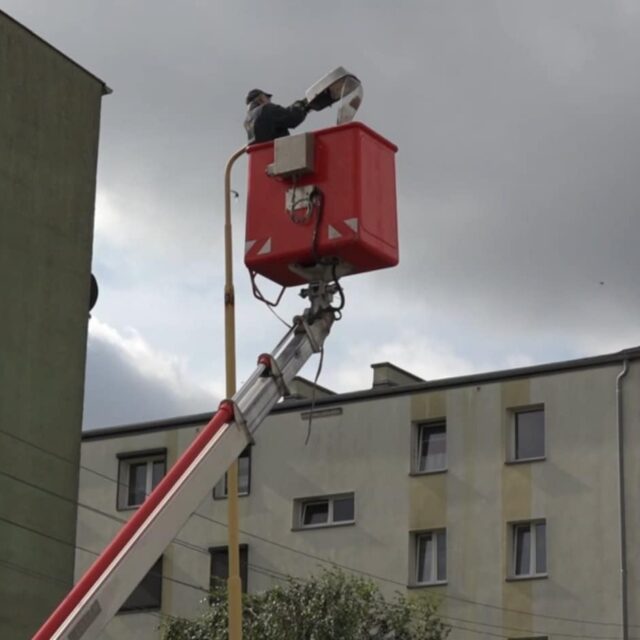 Elektryk wymieniający oprawę oświetlenia na słupie.
