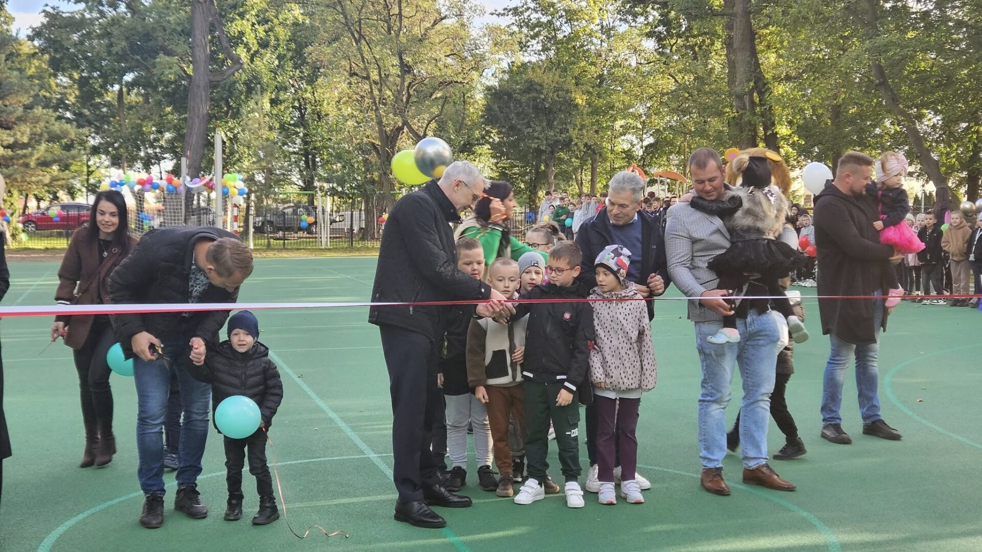 Przecinanie wstęgi przez dorosłe osoby i dzieci na otwarciu wielofunkcyjnego boiska sportowego.