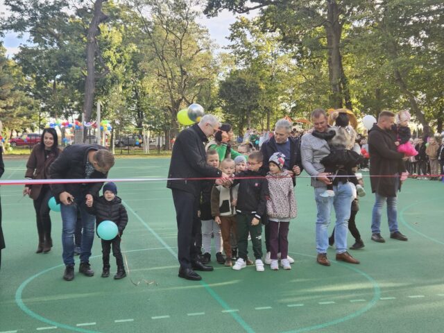 Przecinanie wstęgi przez dorosłe osoby i dzieci na otwarciu wielofunkcyjnego boiska sportowego.