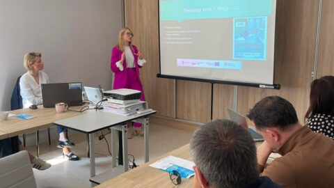 Na zdjęciu kobieta prezentująca prezentacje na ekranie przed osobami siedzącymi przy stołach konferencyjnych.