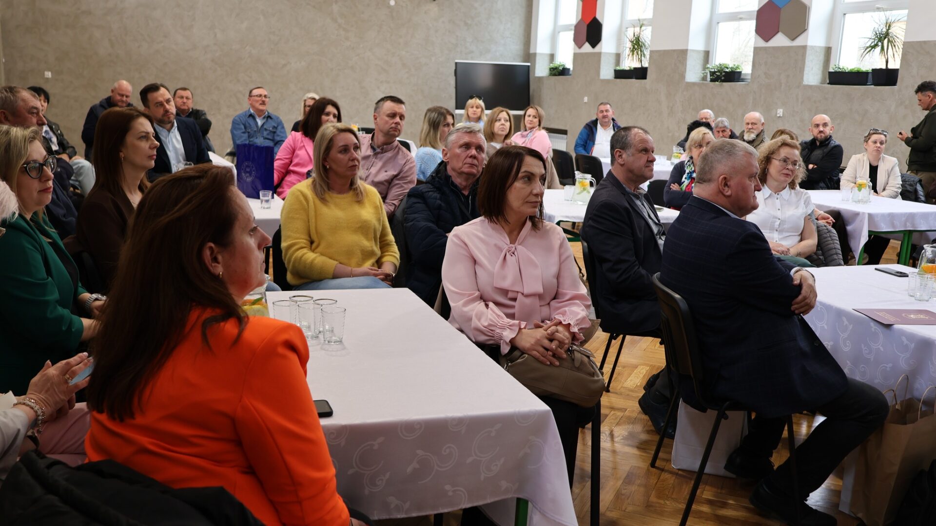 Liczna grupa osób siedząca przy stolikach i słuchająca osoby przemawiającej .