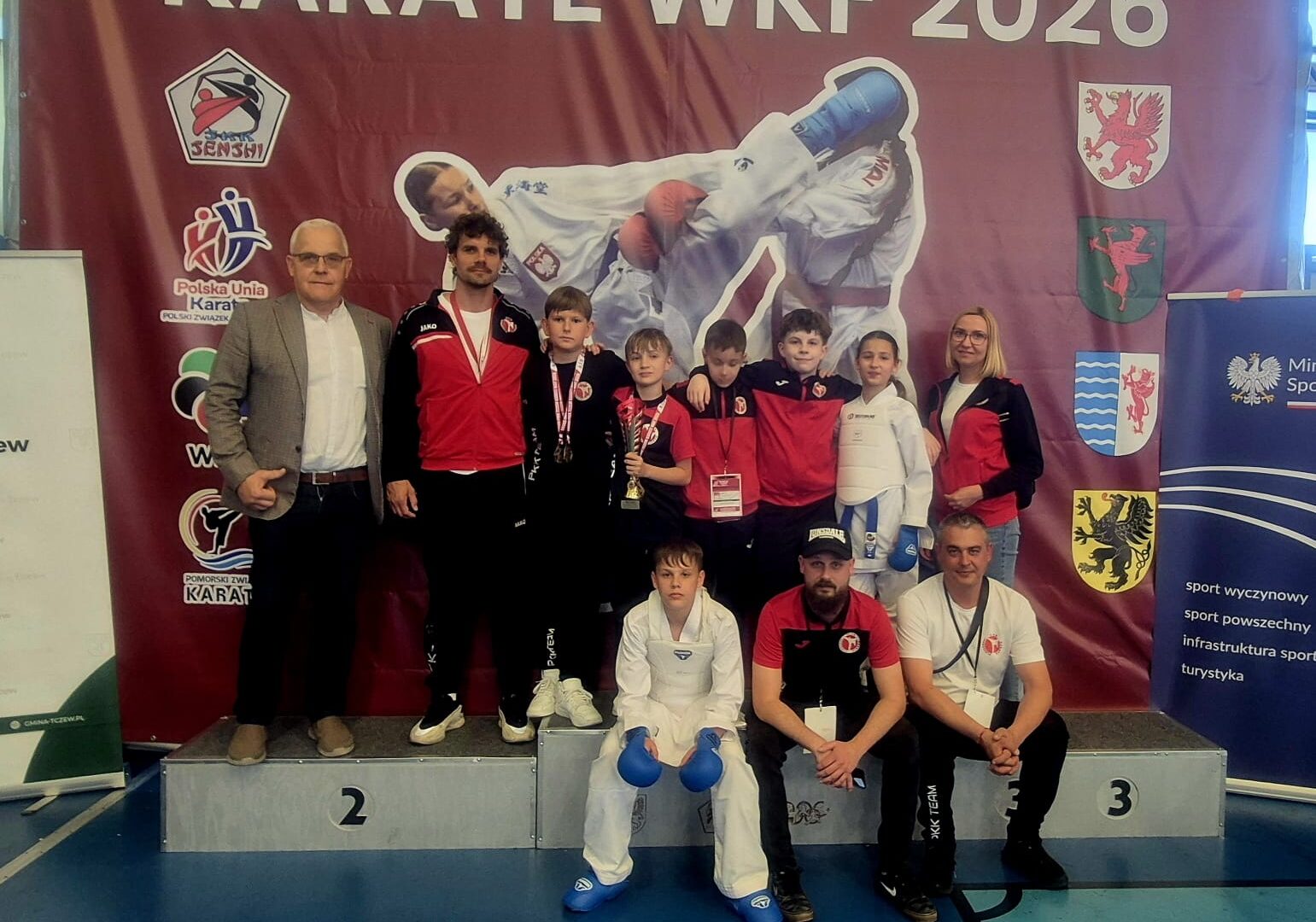 Pleszewska drużyna karate z trenerami na ściance i podium zawodów karate w Tczewie.